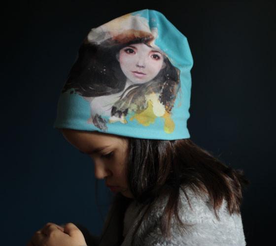 Child wearing a teal beanie with abstract portrait art by Élise Charette on her head