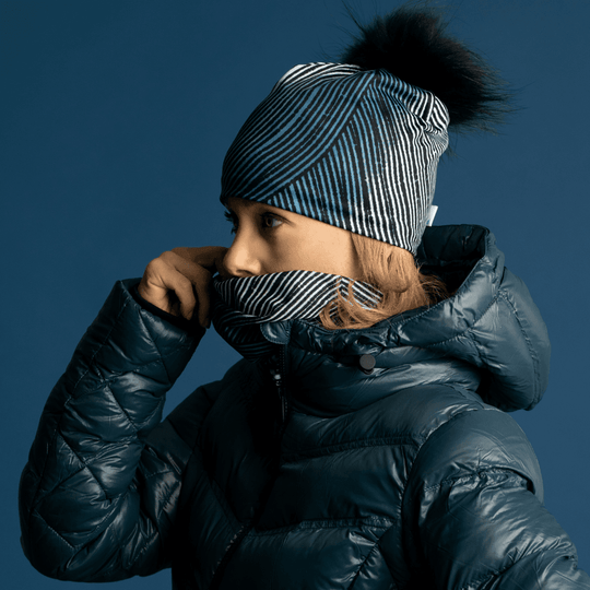 Side profile of a woman wearing a blue winter jacket, abstract patterned beanie, and neck gaiter pulled up to her nose.