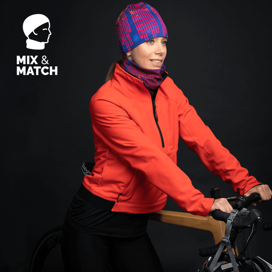 Woman wearing a blue and pink patterned beanie and matching neck tube with an orange jacket, standing by a bicycle.