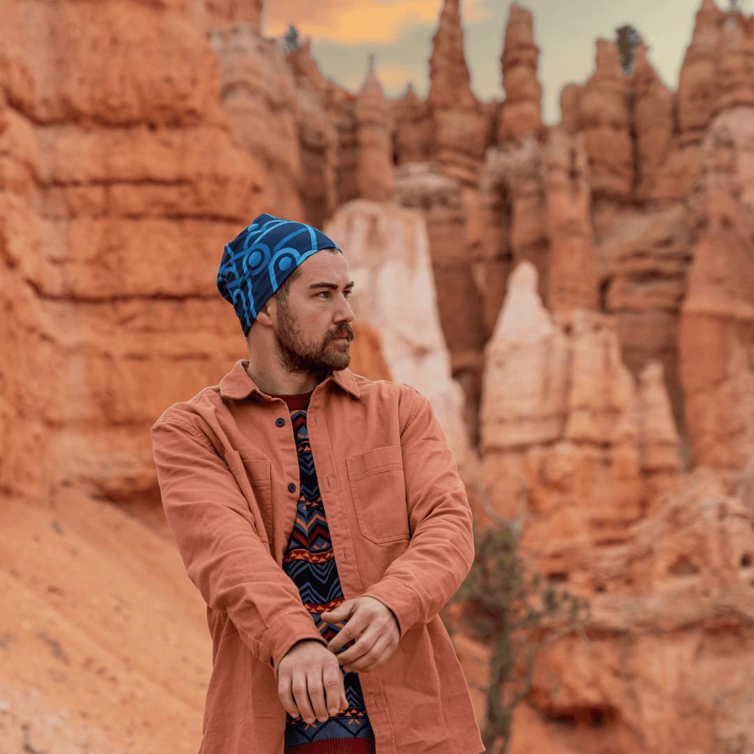 Man with a beard wearing a salmon-colored shirt and patterned beanie, standing in a desert landscape.