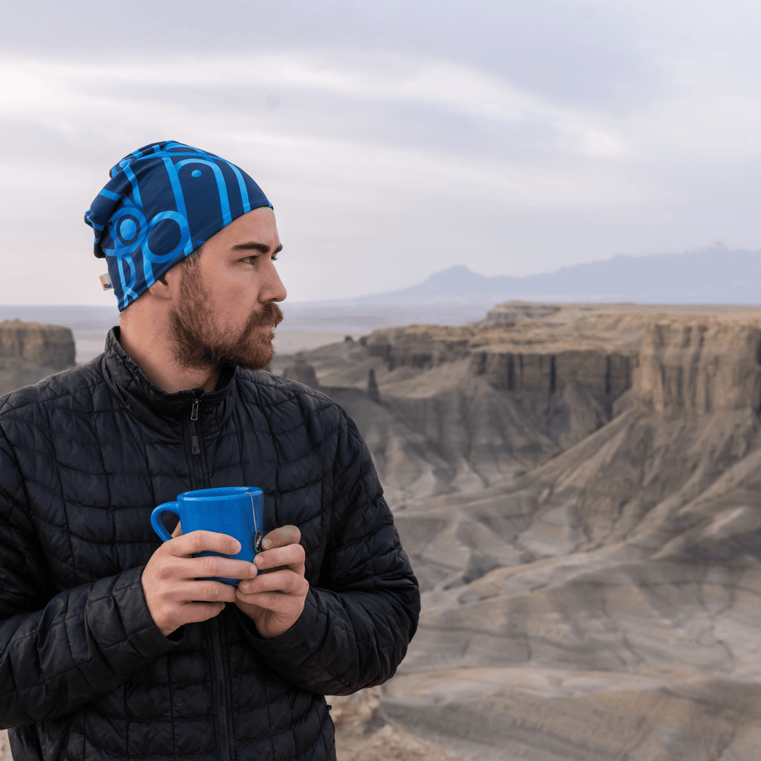 Man wearing a black puffer jacket and a blue patterned beanie, holding a blue mug in a rocky landscape.