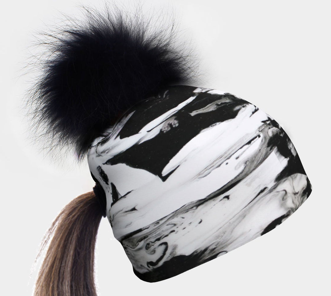 Black and white marble pattern beanie with a large black pompom on top and a ponytail extending through a hole at the back.