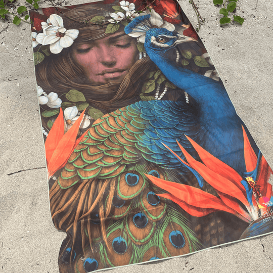 The Chrysalide artistic beach towel laid out on a sandy beach, showcasing its vibrant peacock and floral design.