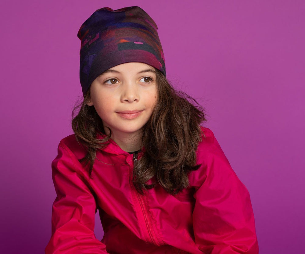 A young girl wearing the City Beanie, which displays a vibrant abstract print, against a solid purple backdrop.