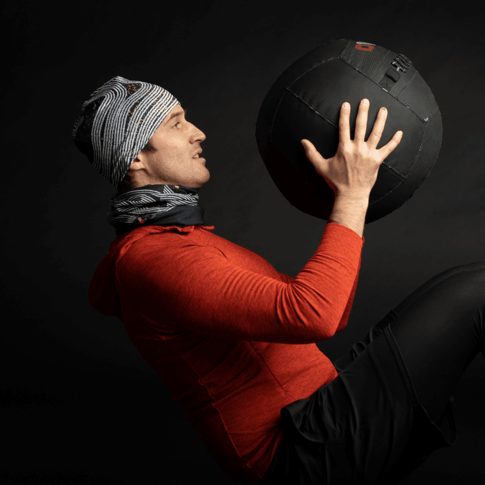 A man in athletic wear wearing a Dark Steel beanie and neck gaiter while lifting a medicine ball.