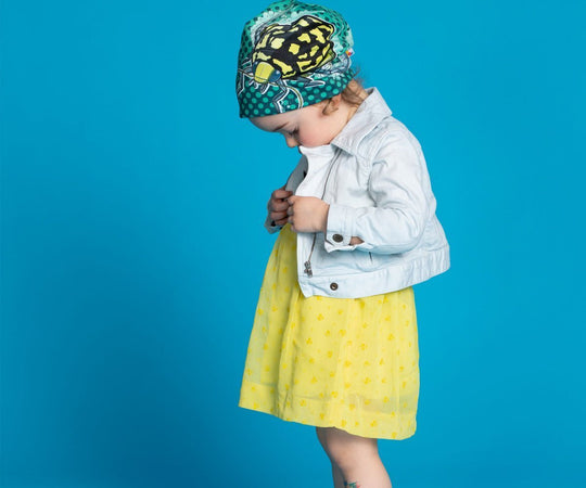 A young child wearing the Diving Beetle Beanie with a light-coloured jacket and yellow dress, looking down.