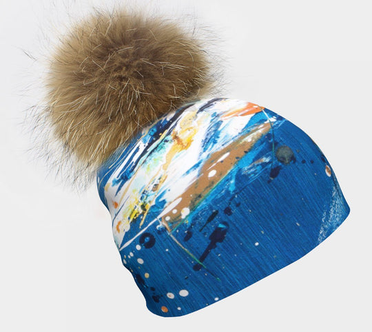 Blue beanie with abstract colourful paint splatters and a large fluffy brown pompom.