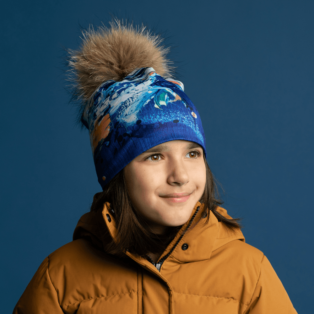 Young girl wearing the blue abstract art beanie with a fluffy pompom and a yellow jacket.