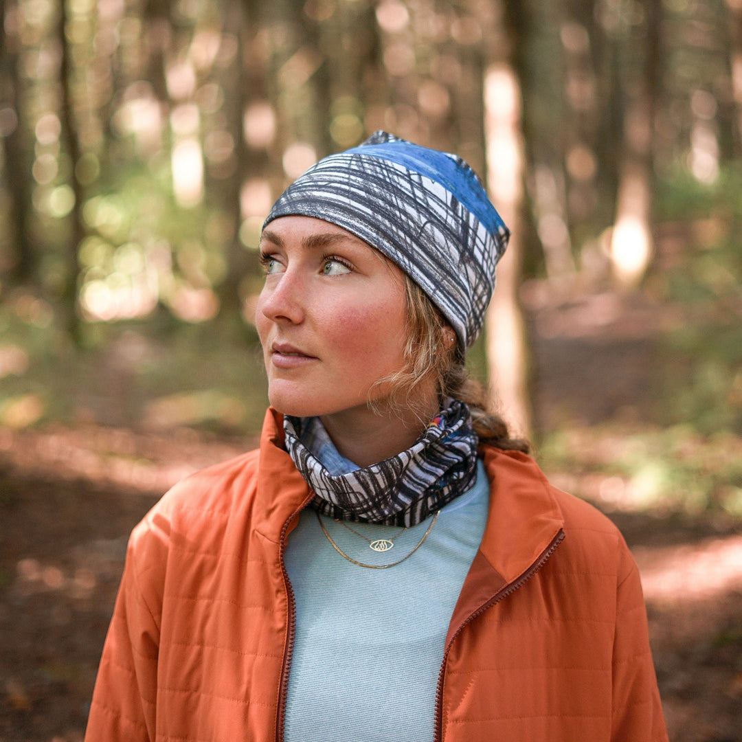 A woman wearing the Expressive Sketch Beanie and neck tube in a wooded area, looking thoughtfully to the side.