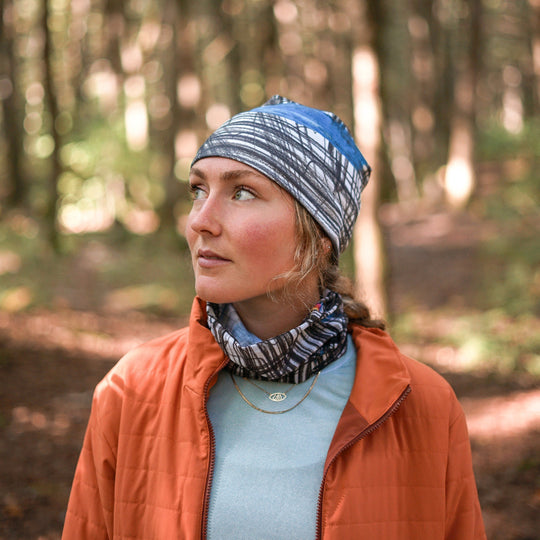 A woman wearing the Expressive Sketch Beanie and neck tube in a wooded area, looking thoughtfully to the side.