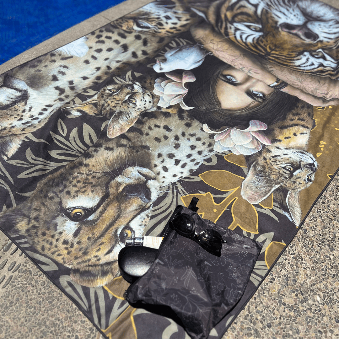 The Felines XL artistic towel laid out by a pool with sunglasses and a pouch.