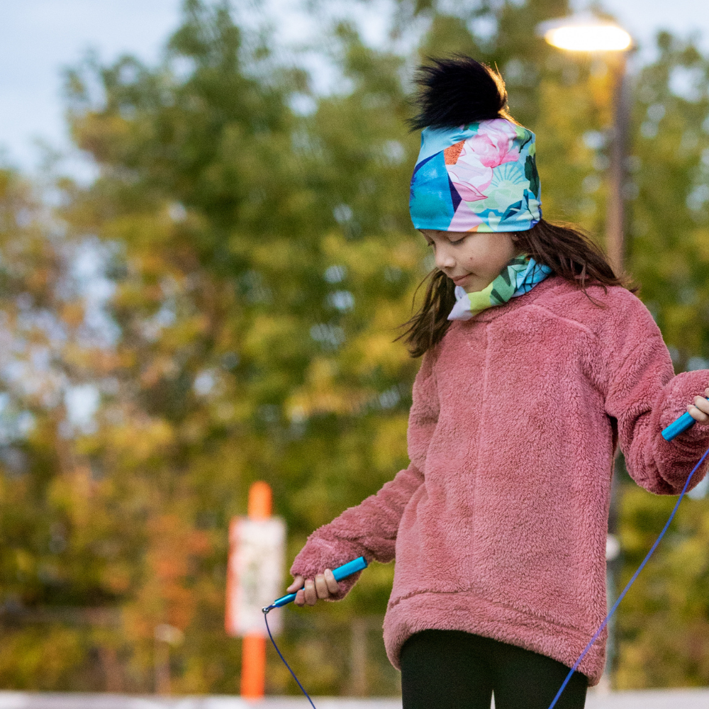 A young girl wearing a matching beanie and scarf, jumping rope outdoors.