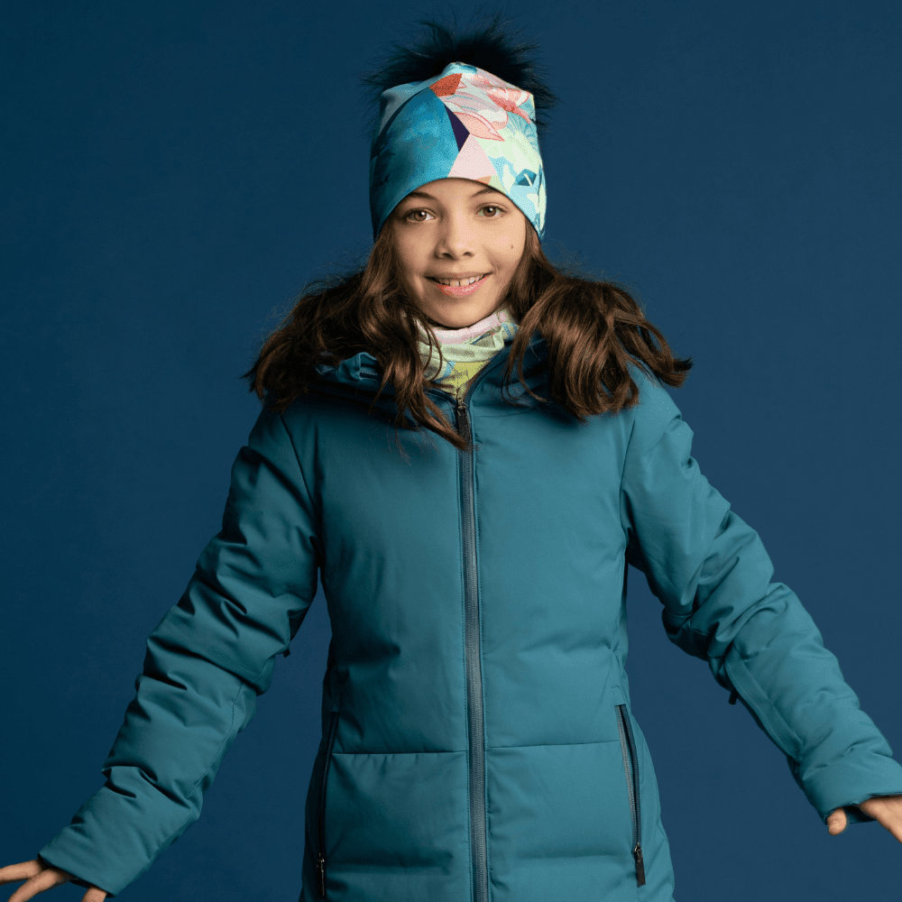 A young girl wearing a matching beanie and scarf, dressed in a teal winter jacket, smiling against a blue background.