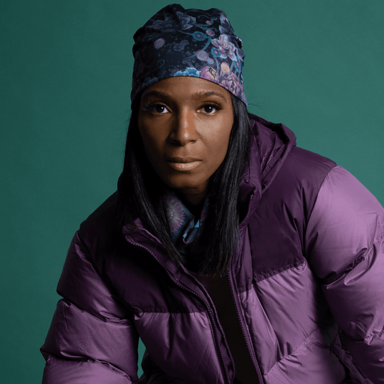 A woman wearing a purple puffer jacket and a floral beanie, with the matching neck tube visible.