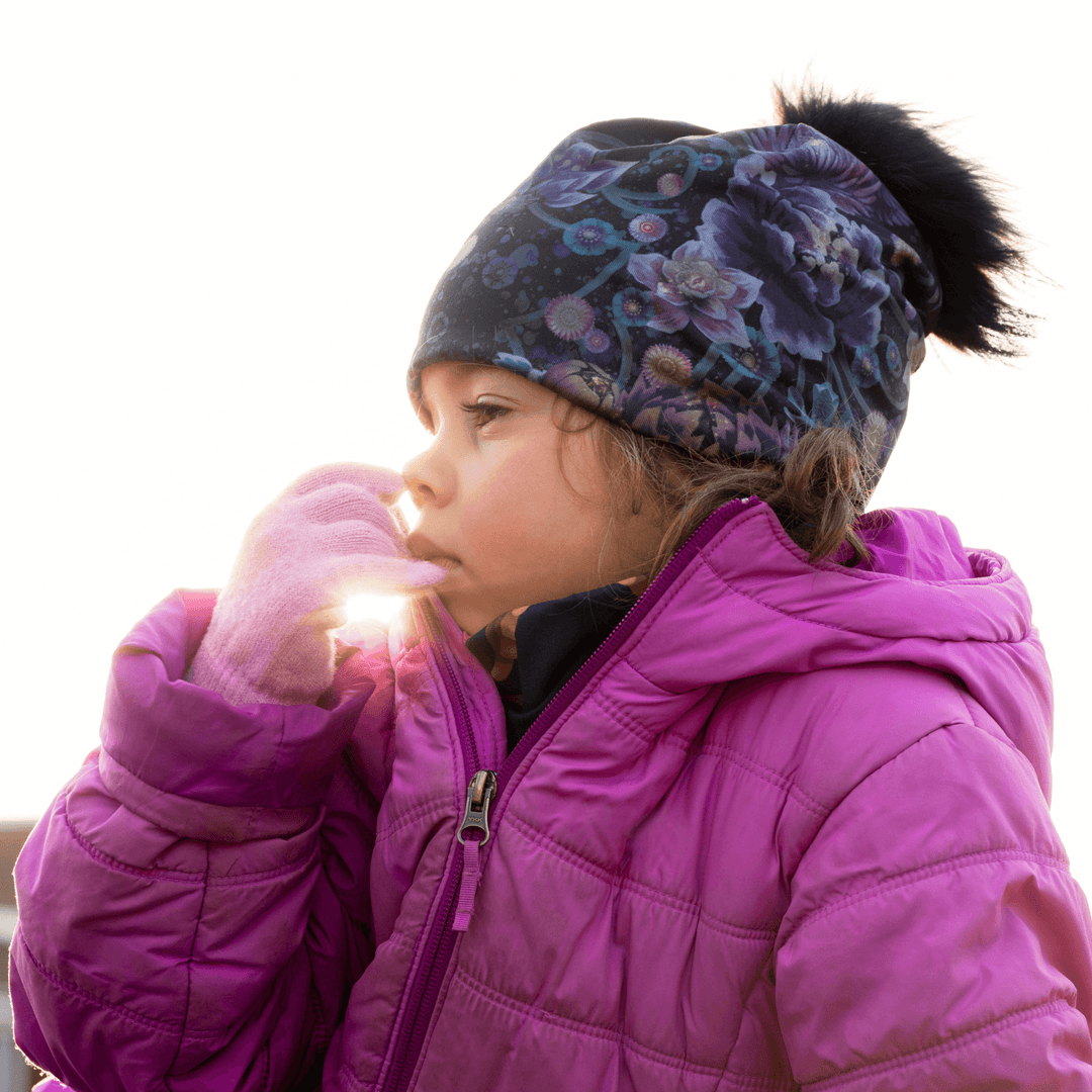 Young child wearing the Flora Pompom Beanie and a pink jacket, with the sun shining brightly behind them.