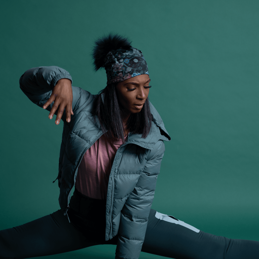 Woman wearing the Flora Pompom Beanie and a grey puffer jacket in a wide yoga split.