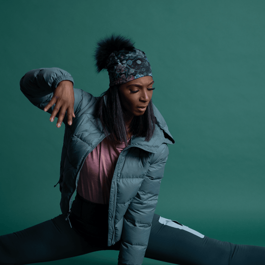 Woman wearing the Flora Pompom Beanie and a grey puffer jacket in a wide yoga split.