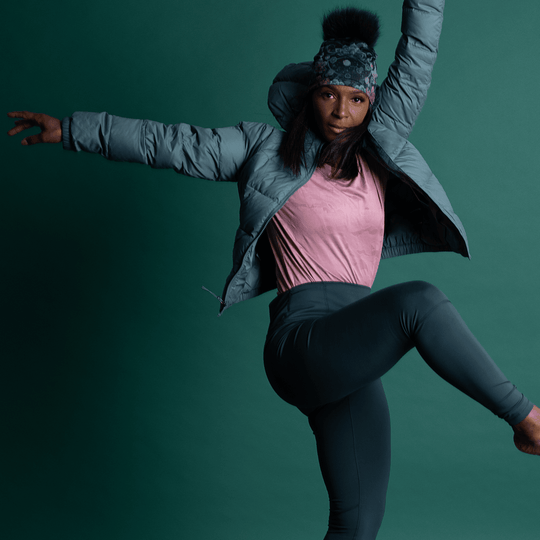 Woman in a grey puffer jacket and dark leggings in a mid-air yoga pose, wearing the Flora Pompom Beanie.
