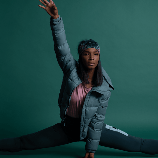 Woman in a grey puffer jacket and dark leggings performing a dynamic yoga pose, wearing the Flora Pompom Beanie.