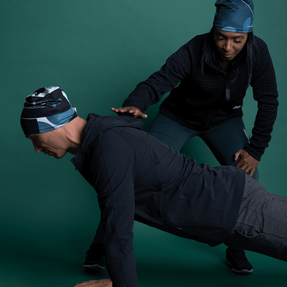 A man and woman wearing beanies outdoors; the man is doing a plank while the woman assists.