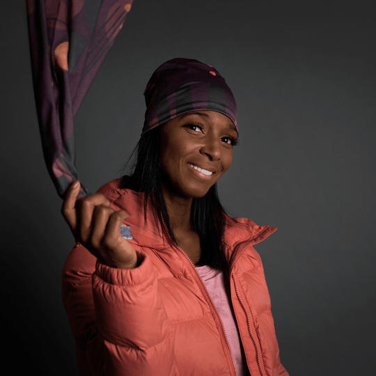 A smiling woman in a coral puffer jacket holds a purple geometric patterned bandana, looking towards the camera.
