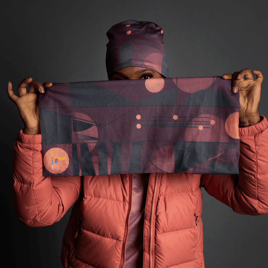 A woman in a coral puffer jacket holds up a unfolded purple geometric patterned bandana.