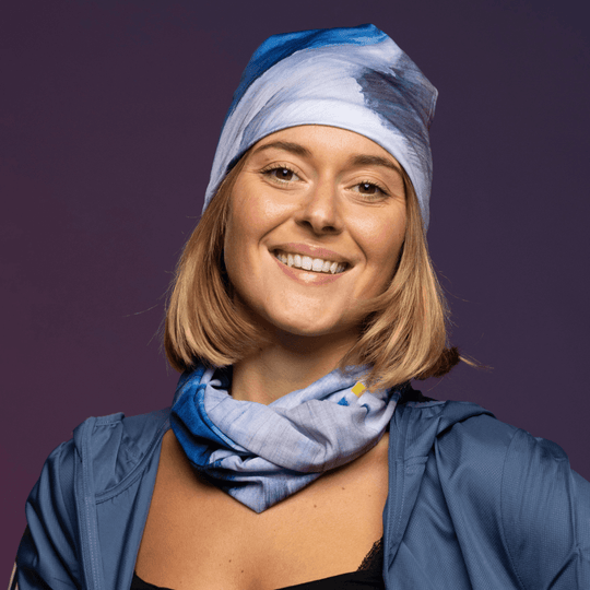 A smiling woman wears a blue jacket and a blue bandana as a neck gaiter, with another bandana tied as a beanie.