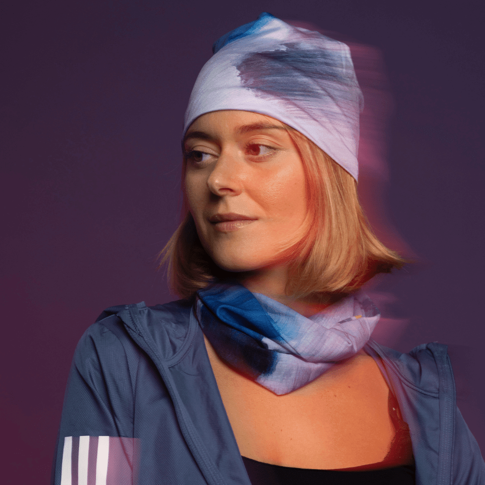 A woman looks to the side, wearing a blue jacket with a bandana tied as a beanie and another as a neck gaiter, with a red-orange glow.