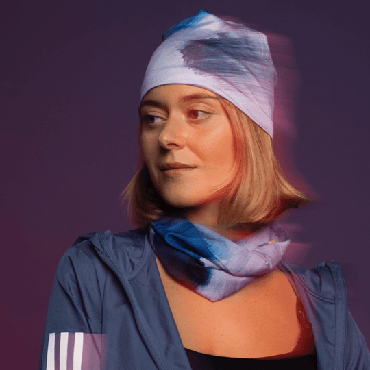A woman looks to the side, wearing a blue jacket with a bandana tied as a beanie and another as a neck gaiter, with a red-orange glow.