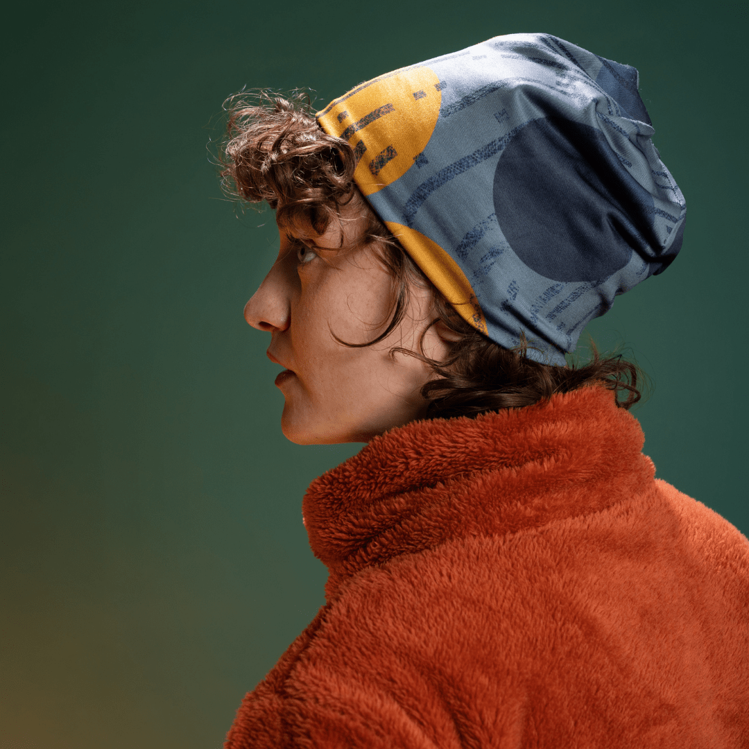 Teen model wearing the grey and gold art beanie with a rust-orange fleece coat, photographed in profile against a green background