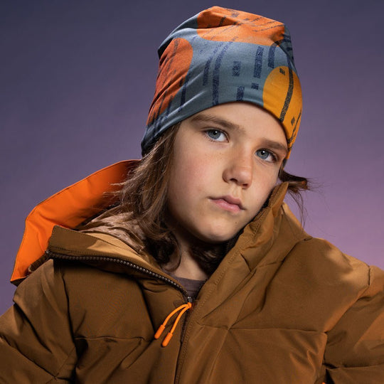 Child wearing the orange and grey art beanie loosely styled with a brown winter puffer jacket, purple studio backdrop