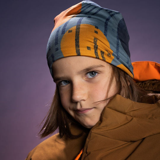 Child in brown puffer jacket looking directly at camera wearing the vibrant orange movement art beanie, purple studio background