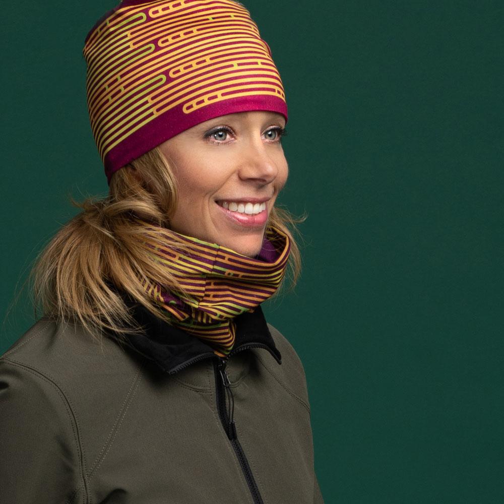 Woman smiling and wearing a matching magenta beanie and neck gaiter, with a green jacket.