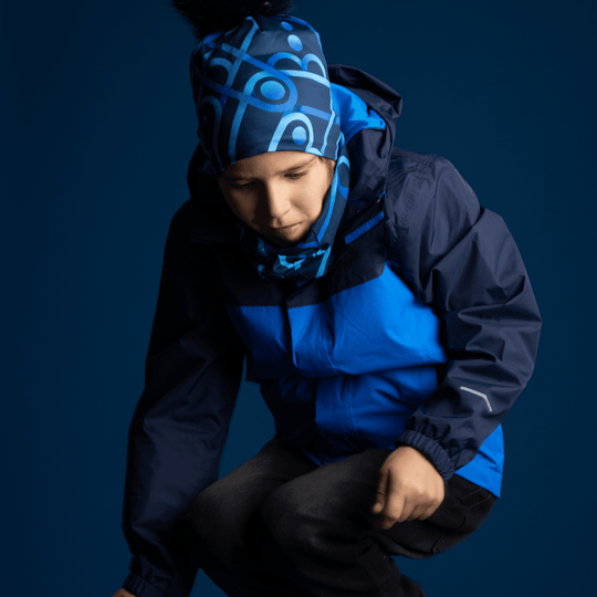 A young boy wearing a blue jacket, Mechanic Blue beanie, and matching neck gaiter, posing in a dark blue studio setting.