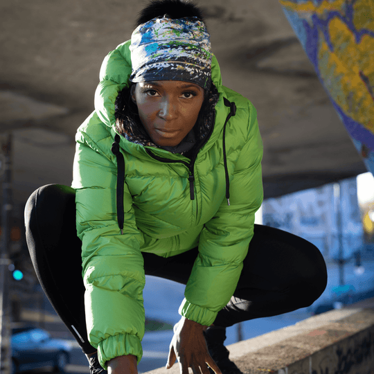 Woman in a squatting pose, wearing the Meduse Beanie with abstract art and a green puffer jacket.