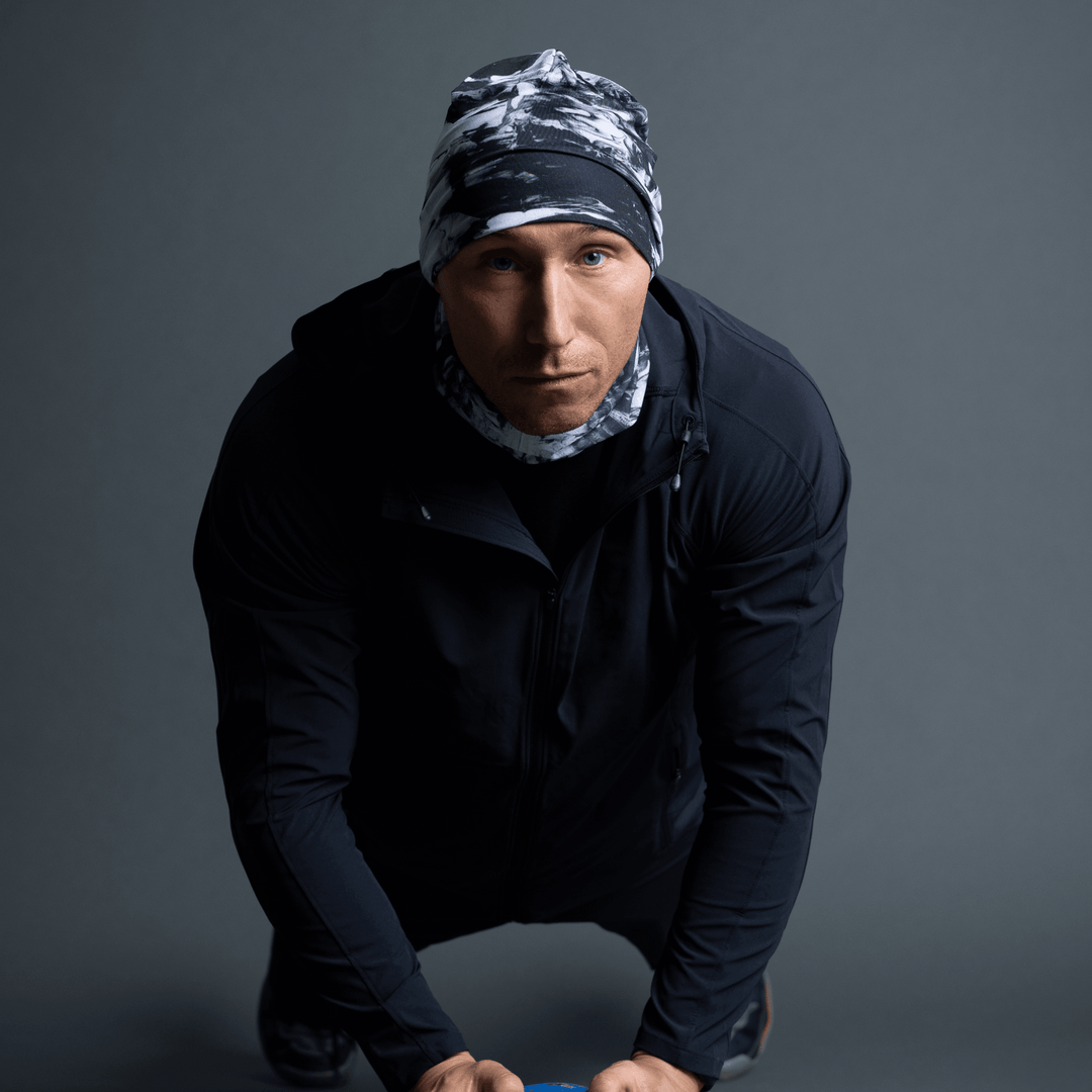 Man in a black jacket wearing a black and white abstract pattern beanie and neck gaiter, in a push-up pose.