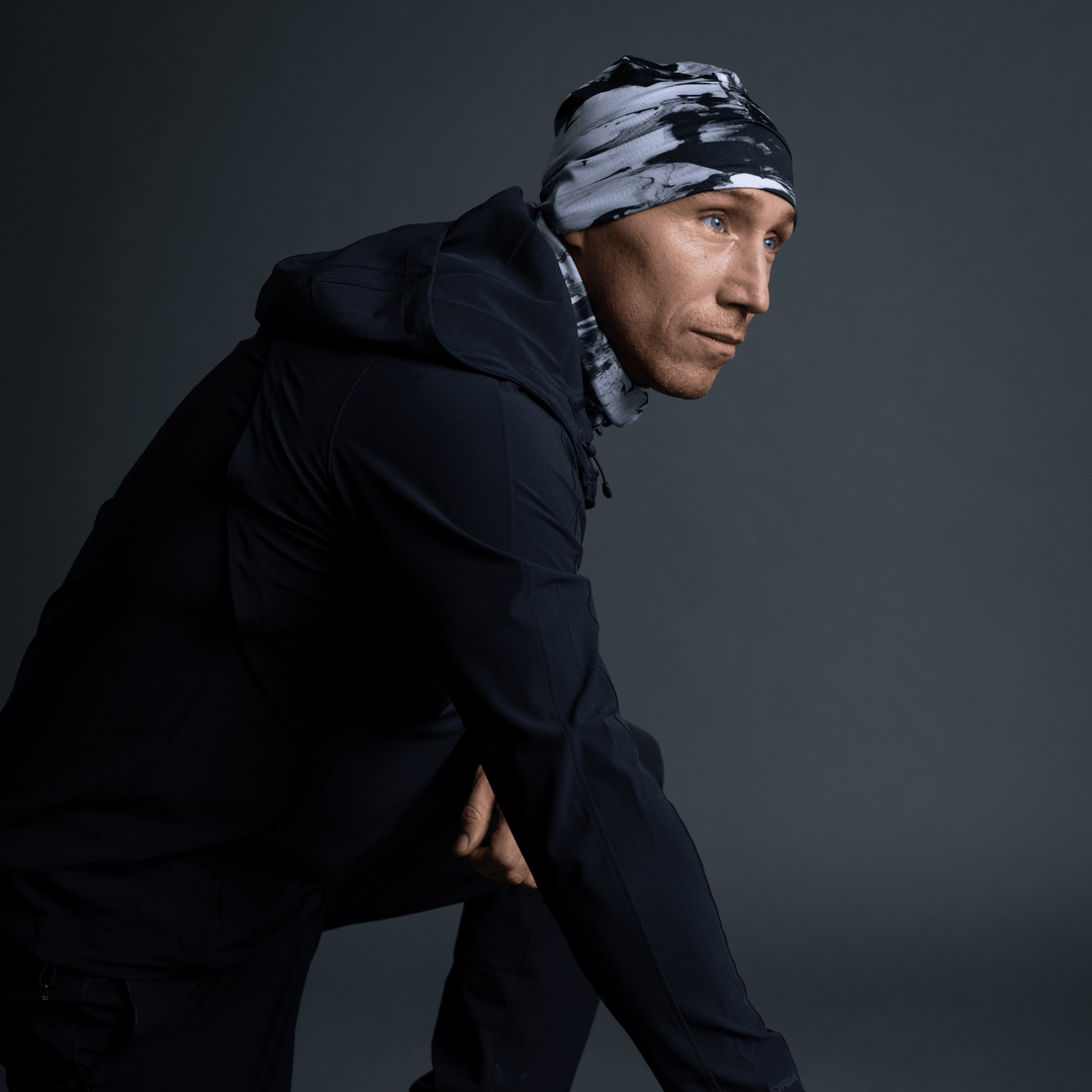 Man in a black jacket and black and white abstract pattern beanie and neck gaiter, in a crouched pose.