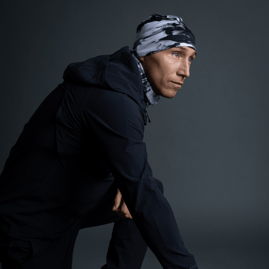 Man in a black jacket and black and white abstract pattern beanie and neck gaiter, in a crouched pose.