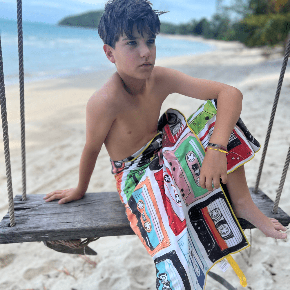 Child on a wooden swing on a beach, wrapped in the colorful side of the Mixtapes beach towel.