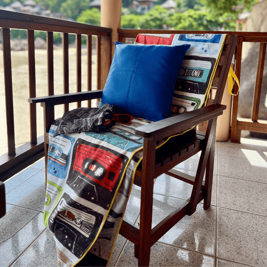 Mixtapes beach towel draped over a wooden chair on a balcony, alongside a blue pillow and sunglasses.