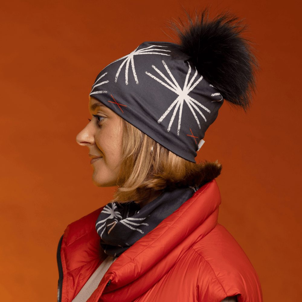 Side profile of a woman wearing the black MotifX beanie with black pompom and a matching scarf, over an orange background.