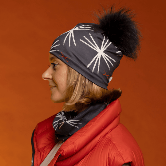 Side profile of a woman wearing the black MotifX beanie with black pompom and a matching scarf, over an orange background.