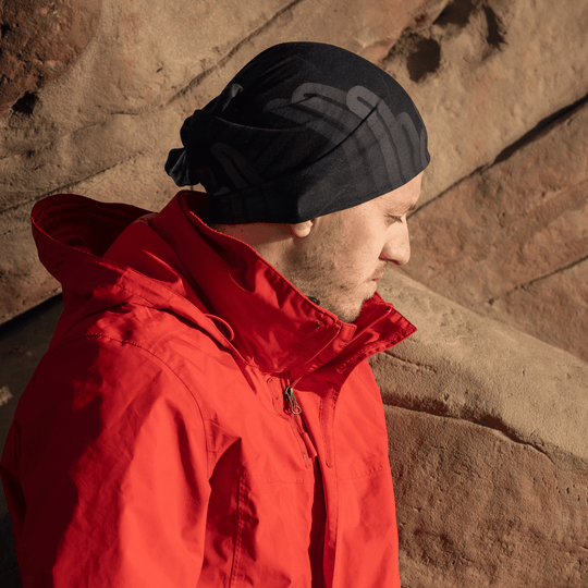 A man wearing a red jacket and a black multifunction tube as a bandana, standing against a rocky backdrop.