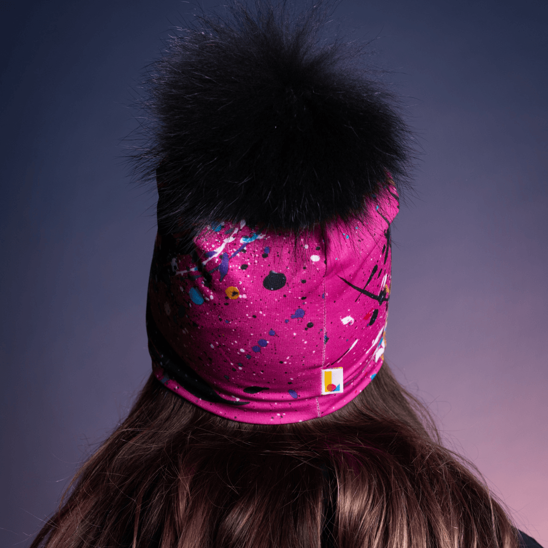 Rear view of a pink beanie with black abstract art splatter and a black pompom, showing the back of the head.