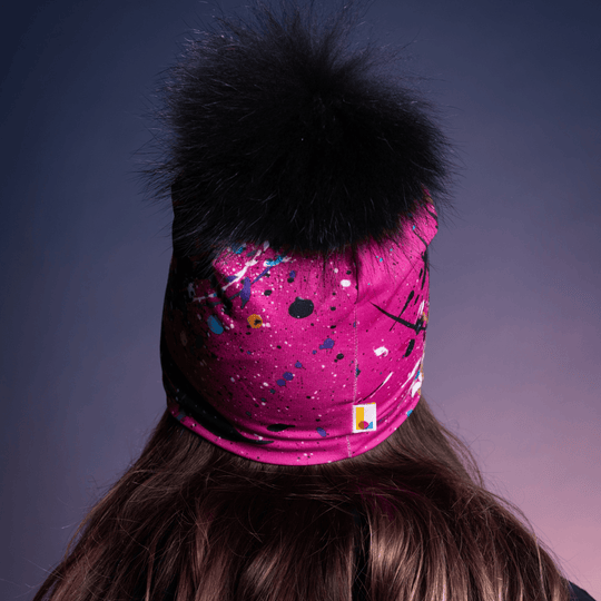 Rear view of a pink beanie with black abstract art splatter and a black pompom, showing the back of the head.
