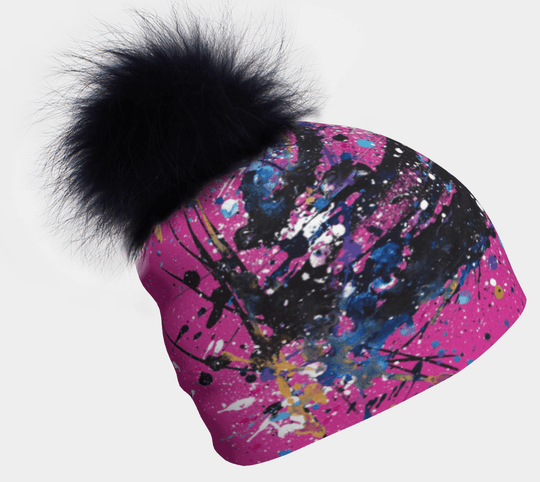 Pink beanie with abstract art splatter in blue, purple, black, and yellow, topped with a large fluffy black pompom.