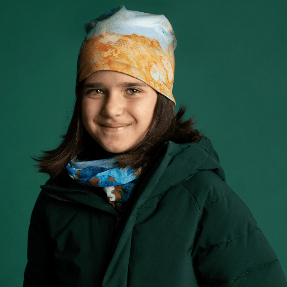 A young girl wearing a green winter coat, a blue multifunctional tube around her neck, and a matching beanie.