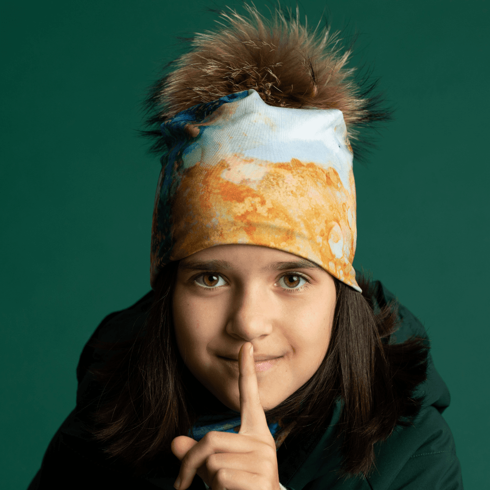 A young girl wearing the 'Ocean Marble' beanie and scarf smiles directly at the camera, with her finger to her lips.