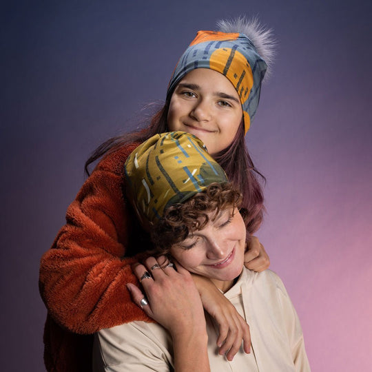 Two young people wearing bold orange-and-blue patterned beanies in a warm studio portrait, showcasing the wearable-art style