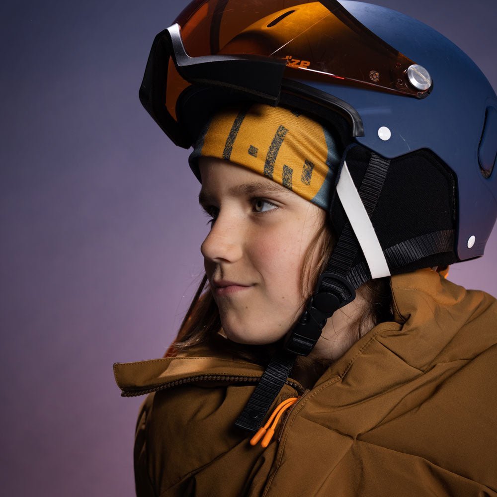 Side profile of a child in ski gear wearing the abstract yellow-and-teal beanie beneath a blue ski helmet, showing a slim helmet-ready fit